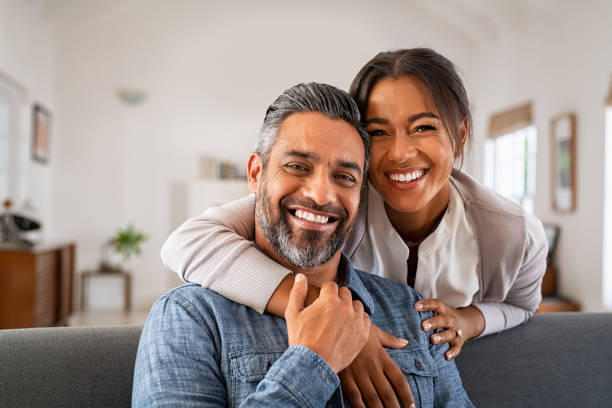 Portrait of multiethnic couple embracing and looking at camera sitting on sofa. Smiling african american woman hugging mid adult man sitting on couch from behind at home. Happy mature mixed race couple laughing at home.